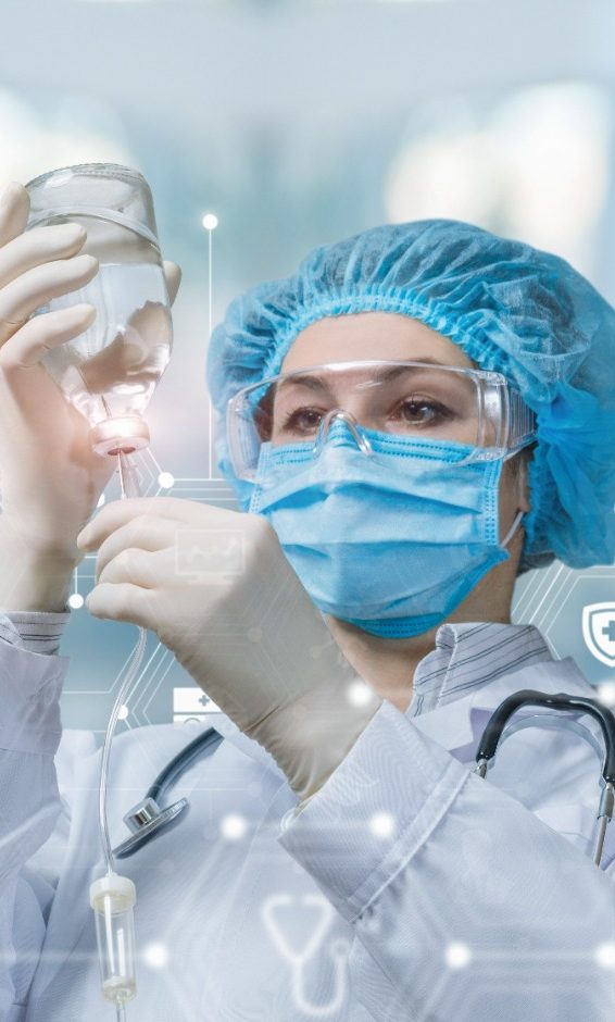Ärztin bereitet in Schutzkleidung das System für Infusionen vor, während im Hintergrund ein verschwommenes Bild zu sehen ist