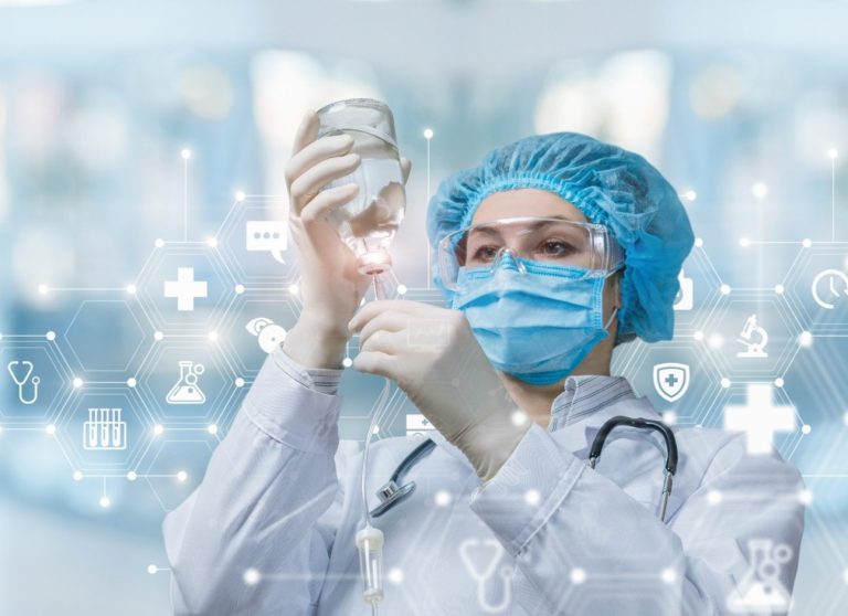 Ärztin bereitet in Schutzkleidung das System für Infusionen vor, während im Hintergrund ein verschwommenes Bild zu sehen ist